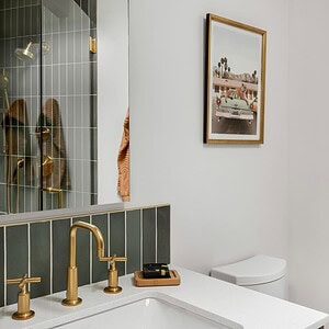 A modern bathroom with a white countertop, gold faucet, wall-mounted mirror, framed artwork, and a toilet. The backsplash features vertical dark tiles accented with a touch of Eichler Orange.