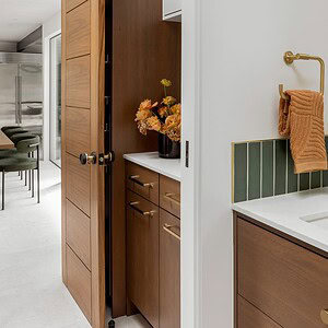 Modern kitchen with wooden cabinets and door. Countertop adorned with a floral arrangement beside a sink with a green tile backsplash, Eichler Orange accents, and a brown towel on a gold holder.