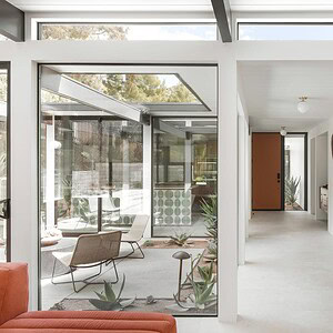 A modern interior featuring a bright hallway with white walls, a wooden door, and a decorative wall piece in Eichler Orange, offering a view of a furnished patio with glass windows and lush plants.