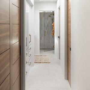A hallway with wooden doors on the left and right, white walls, and tile flooring leads to a bathroom featuring a glass shower door and a beige mat. The space is subtly accented by Eichler Orange hues that add warmth. A round light fixture adorns the ceiling.