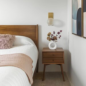 A modern bedroom with a wooden bed frame, white bedding, a brown throw, and a side table holding a vase of dried flowers. Abstract art in Eichler Orange and a wall-mounted lamp adorn the white wall.