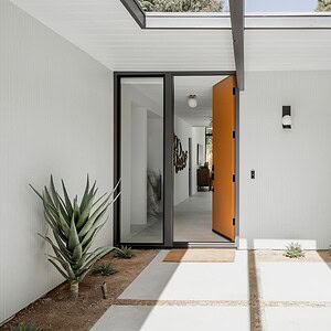 A modern home entrance with a partially open Eichler Orange door, potted plants, skylight, and a vintage orange and white car parked in a minimalist driveway.