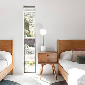 A bedroom with two wooden twin beds, each adorned with white linens and neutral-colored pillows. A small wooden nightstand with a lamp and Eichler Orange decor sits between the beds, perfectly positioned in front of a narrow window.