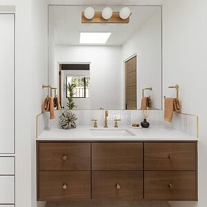 Modern bathroom with a wide mirror above a wooden vanity, white cabinets with brass handles to the left, and brass fixtures including a wall-mounted faucet. Neutral decor accents are complemented by pops of Eichler Orange for an added touch of mid-century flair.