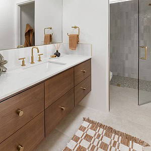 A modern bathroom features a large mirror, a double-sink wooden vanity with brass fixtures, and a checkered rug. The space is complemented by an Eichler Orange accent wall and a glass-door shower with light-colored tiles.