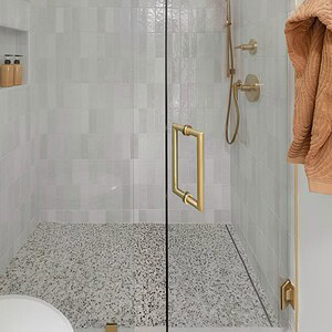 A modern shower enclosure with glass doors, brass fixtures, a rainfall showerhead, hand-held shower, and built-in shelves. The shower walls have white tiles contrasted by an accent of Eichler Orange, and the floor features speckled stone tiles.