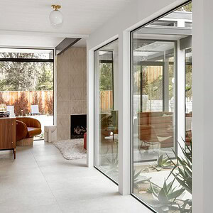 A modern minimalist interior with large windows and an atrium, showcasing an open living space with white walls, light-colored flooring, mid-century furniture in Eichler Orange, and plants.