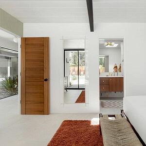 A modern white bedroom with an open wooden door, mirrors, and a view into the bathroom featuring a wooden vanity. The bed with a white duvet and woven bench is accented by pops of Eichler Orange, adding a mid-century flair.