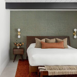Modern bedroom with a mid-century style featuring a double bed, accent pillows, matching nightstands with wall-mounted lamps, and a decorative green geometric accent wall. An Eichler Orange rug sits below.