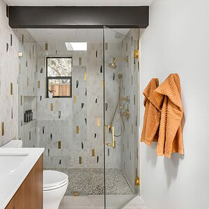 Modern bathroom featuring a glass-enclosed shower, white vanity with gold fixtures, and two Eichler Orange towels on a wall hook. The shower has patterned tiles with gold, gray, and beige accents.