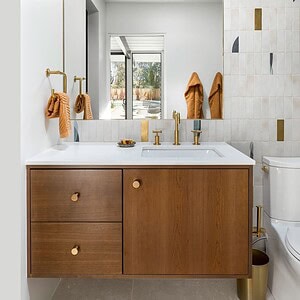 Modern bathroom with a wooden vanity, gold fixtures, a large mirror, white countertop, and beige textured tiles. Towels hang on gold hooks and natural light enters through a window bathed in soft hues reminiscent of Eichler Orange.