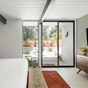 A modern bedroom with a large bed, an Eichler Orange bench at the foot, a dresser, a wall-mounted TV, and sliding glass doors leading to an outdoor patio with chairs.