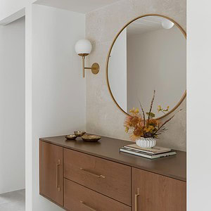 Entrance hallway with a modern, minimalist design featuring a wooden console table, a round mirror, and a vase with flowers. The hallway has an Eichler Orange door and a white wall with a sconce light.