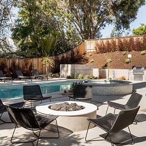 The outdoor patio features a pool, a hot tub, and a fire pit surrounded by black chairs. Lounge chairs beside the pool add relaxation, all set against a backdrop of trees and a wooden fence. The patio's accents in Eichler Orange bring vibrancy to this serene setting.