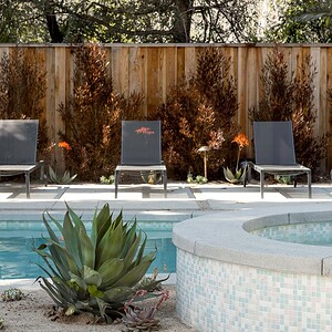 Four empty lounge chairs line the edge of an outdoor swimming pool with a hot tub in the foreground, surrounded by a wooden fence and greenery. The scene is accentuated by touches of Eichler Orange, adding a vibrant contrast to the serene setting.
