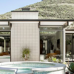 A modern building with large glass windows, displaying a hint of Eichler Orange, stands behind a round tiled hot tub connected to a swimming pool, all set against a backdrop of green hills.
