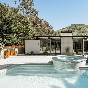 Modern home with large windows and a backyard featuring a swimming pool, hot tub, patio furniture, and surrounded by lush greenery and hills. The design nods to Eichler Orange aesthetics, merging mid-century charm with contemporary comfort.