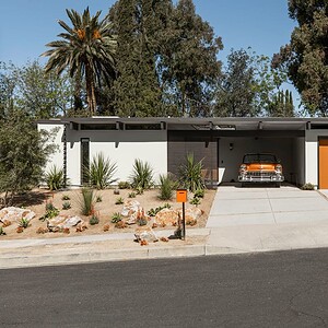 A mid-century modern Eichler house with a flat roof, large windows, and an Eichler orange garage door. A vintage car is parked in the driveway, surrounded by a landscaped yard with desert plants and rocks.
