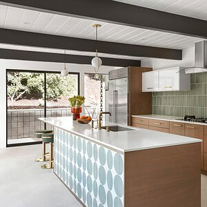 Modern kitchen with a center island featuring blue circular patterns, wooden cabinets, stainless steel appliances, a green tiled backsplash, and large windows flooding the space with natural light— all accentuated by subtle touches of Eichler Orange for a retro flair.