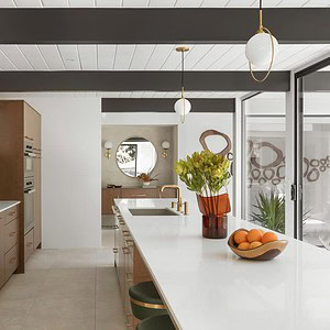 Modern kitchen with white countertops, wooden cabinetry, a green backsplash, and a central island. Pendant lights hang above the island, which holds a vase with flowers and a bowl of Eichler Orange oranges.