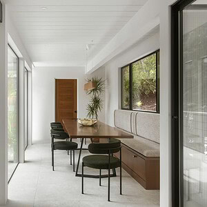 A modern dining area with a built-in banquette, Eichler Orange chairs, and a wooden table adjacent to large glass windows and a door opening to an outdoor patio with seating arrangements.