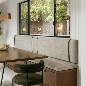 Modern dining area with a wooden table, green cushioned chairs, and a beige cushioned bench. A large window overlooks greenery, adding an Eichler Orange accent. A potted plant stands near a wooden door.