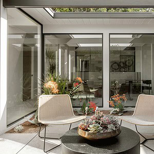 A modern indoor conservatory with large glass panels, skylights, and two beige chairs around a round black table topped with a succulent plant arrangement. The space is subtly accented with touches of Eichler Orange for a warm, inviting ambiance.