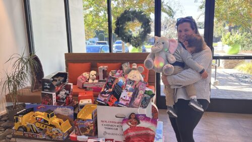 A mom holding a sleepy toddler poses for a picture donating a stuffy to Toys for Tots