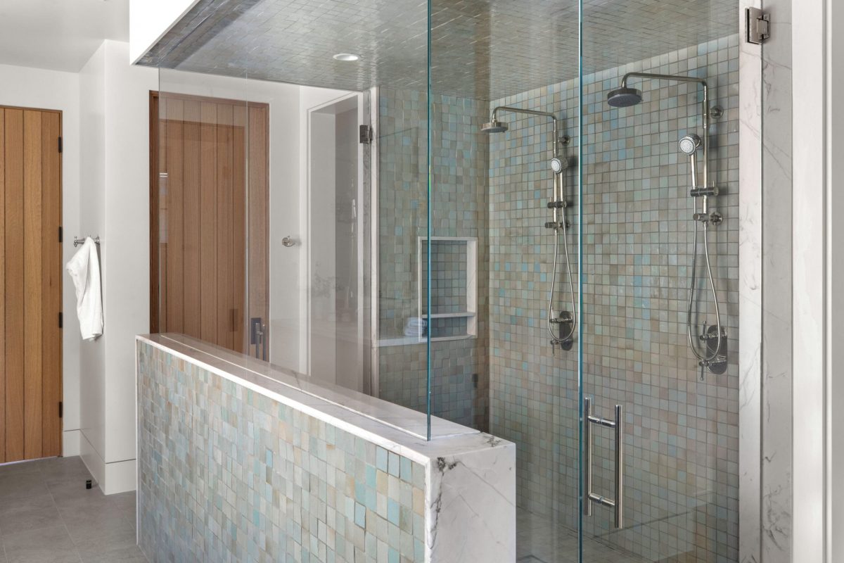 A modern bathroom with a glass-enclosed shower featuring two showerheads, tiled walls, and a raised half-wall partition. A wooden door and towel rack are visible outside the shower.
