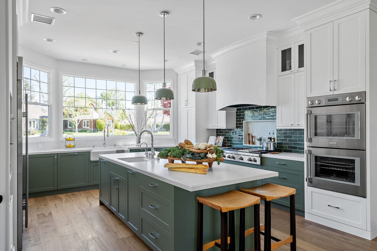 A modern thymeless kitchen with green cabinets, a large center island, three pendant lights, double wall ovens, and a large window providing ample natural light. The island is adorned with a tray of vegetables.
