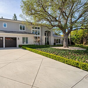 A large, modern two-story house in Westlake Trails features three garage doors, a light-colored exterior, numerous windows, manicured bushes, and an expansive driveway. A large tree stands to the right of the property.