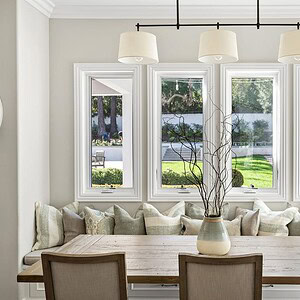 The dining area at Westlake Trails features a wooden table, beige chairs, a cushioned bench with pillows beneath three large windows showing a garden view, and a minimalist chandelier overhead.