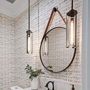 A Westlake Trails modern bathroom features a round mirror, hanging pendant lights, a white countertop, dark wood vanity, and wallpaper adorned with multiple small faces. A potted plant and soap dispenser sit elegantly on the counter.