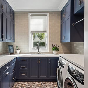 A laundry room in Westlake Trails features navy blue cabinets, a white countertop, a washer and dryer, a window with a shade, beige subway tile backsplash, and potted plants.