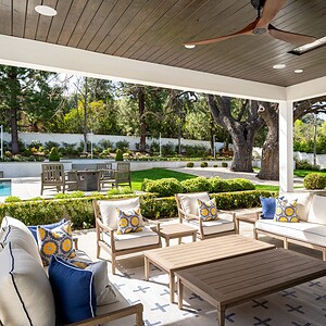 A patio in Westlake Trails featuring cushioned seating with patterned pillows, a wooden coffee table, a ceiling fan, and a view of a landscaped garden, pool, and outdoor dining area with greenery and trees in the background.