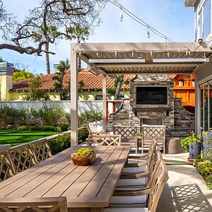 Outdoor patio with a wooden dining table and chairs, a pergola with string lights, and a stone fireplace. Lush greenery and a lawn area are visible in the background, capturing the serene ambiance of Westlake Trails.