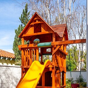 A wooden playhouse with an attached yellow slide and a swing, situated in a backyard with greenery and a white fence in the background, perfect for the idyllic Westlake Trails community.