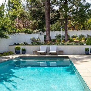 A backyard in Westlake Trails features a swimming pool, two white lounge chairs, potted plants, a garden with lush greenery, and trees in the background. A set of stairs leads up to a raised garden area.