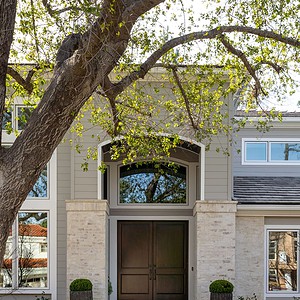 A modern house exterior in Westlake Trails features a large black front door, beige brick façade, large windows, and a tree with green leaves partially shading the entrance.