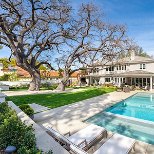 A spacious backyard in the prestigious Westlake Trails features a swimming pool, lounge chairs, outdoor seating, and a well-kept lawn with large trees, adjacent to a two-story gray house under a clear blue sky.