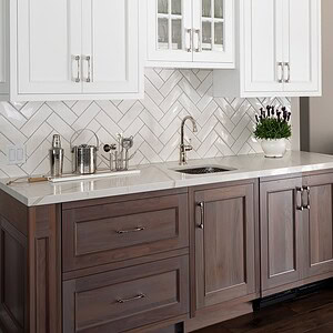 In the heart of Westlake Trails, a kitchen area with upper white cabinets featuring glass panels, lower wooden cabinets, a white herringbone tile backsplash, stainless steel sink and faucet, and a potted plant with purple flowers offers both style and functionality.