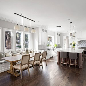 A spacious kitchen in Westlake Trails features white cabinetry, a large island with three stools, a wooden dining table with chairs and bench seating, stainless steel appliances, and large windows providing natural light.