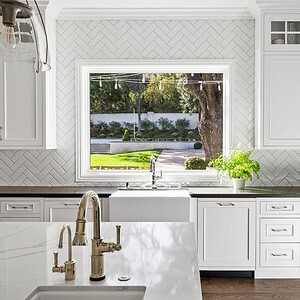 Bright kitchen with white cabinets, a large window above the sink offering a view of the Westlake Trails' lush greenery, and a white herringbone tile backsplash. Marble island with a built-in faucet in the foreground.