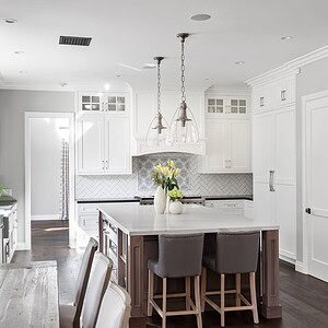 A modern kitchen in Westlake Trails features white cabinets, a kitchen island with two chairs, pendant lights, and stainless-steel appliances. A dining table and a sign on the wall complete this elegant space.