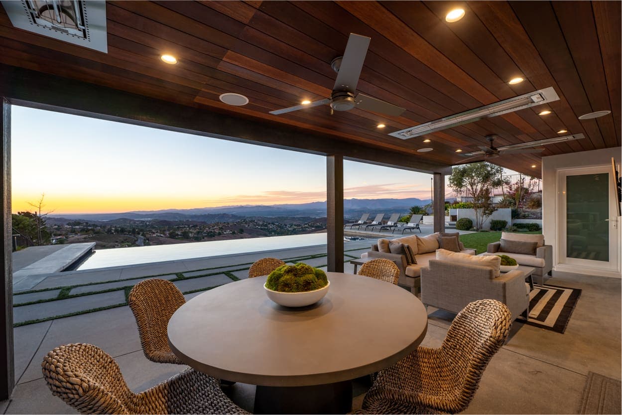 Covered outdoor patio with a circular table and wicker chairs, overlooking a modern pool and lounge chairs. Perfect for your evening projects, the space also offers a scenic view of the sunset and mountains in the background.