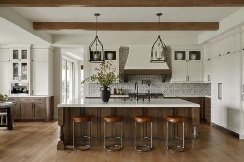 A modern kitchen, truly a Mediterranean marvel, features a large island with four stools, pendant lights, white cabinetry, a patterned backsplash, and wooden ceiling beams.