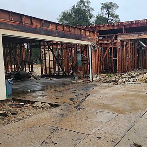 A partially demolished house with exposed wooden framing, rubble scattered on the ground, and a large dumpster nearby—an Organic Modern Home coming soon.