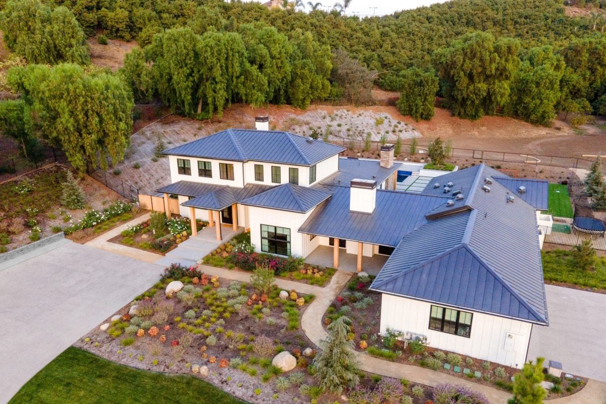 Aerial view of a modern two-story house with a gray metal roof, large windows, landscaped garden, and surrounding trees—showcasing what it really takes to transform a home into a stunning retreat.