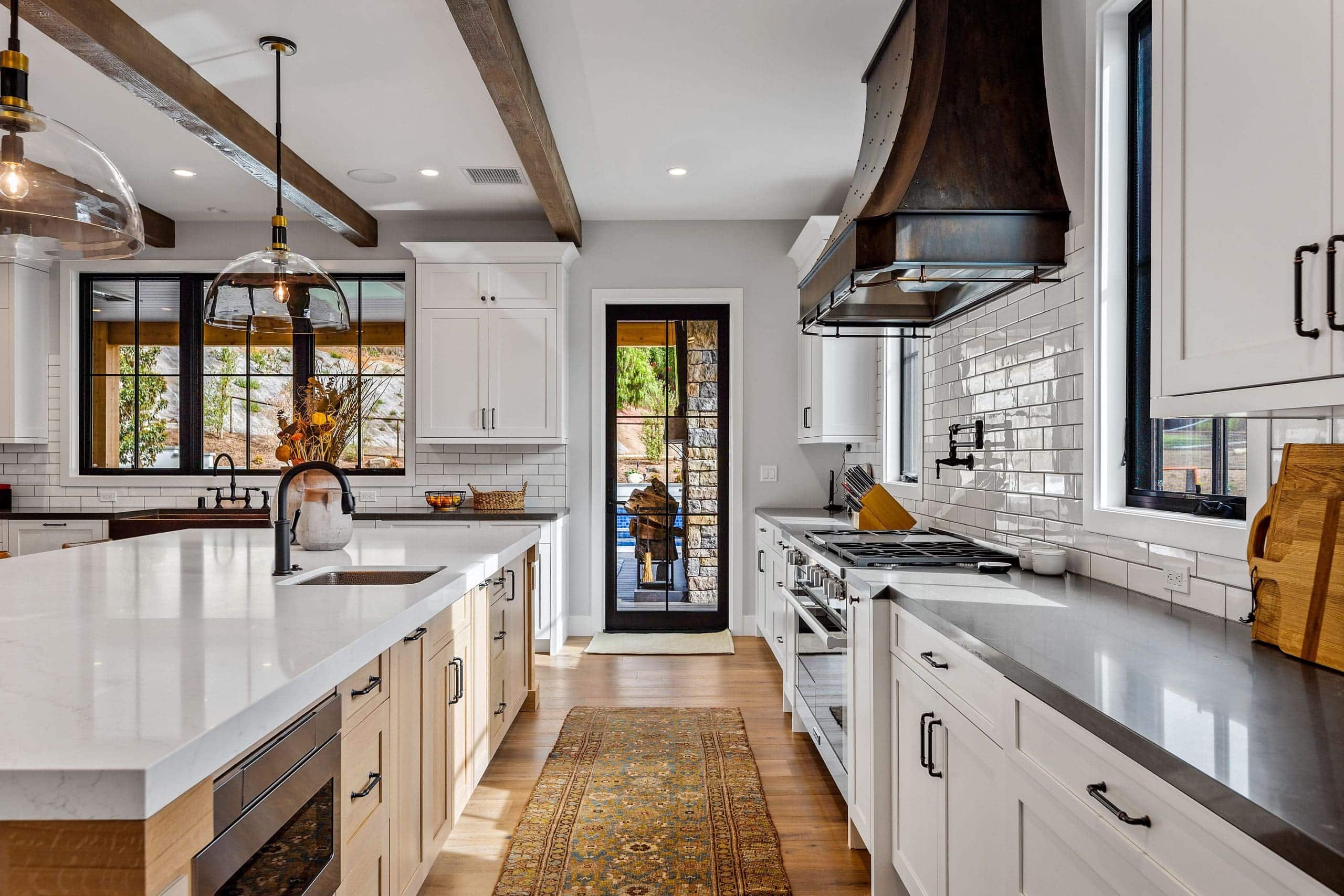 Modern kitchen with white cabinets, large island, stainless steel appliances, wooden beams, and a patterned rug—showcasing what it really takes to transform a home—with a door leading outside.