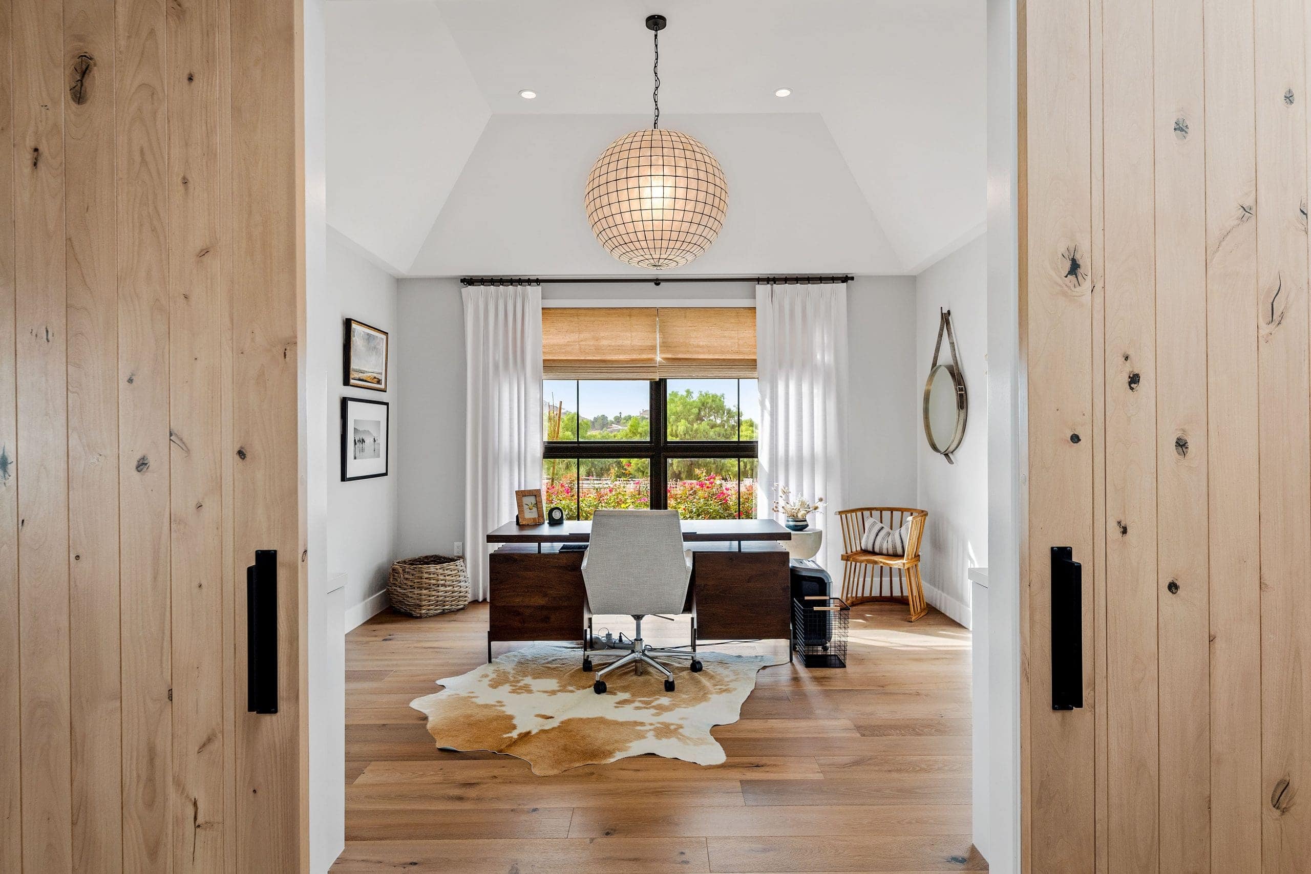 A modern home office with a wooden desk, white chair, animal hide rug, and large window with white curtains—viewed through double wooden doors—shows What It Really Takes To Transform A Home into a stylish and functional space.